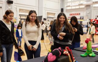 This image shows three Noble Schools students smiling at some art pieces on the table at the Visual Arts Festival. Behind them, you can see other people viewing all the student artwork displayed in the Chicago Bulls College Prep gym.