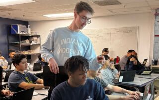 Cameron Forsythe is in his classroom talking to a student, standing beside him and pointing at the computer. They both are looking at the computer.